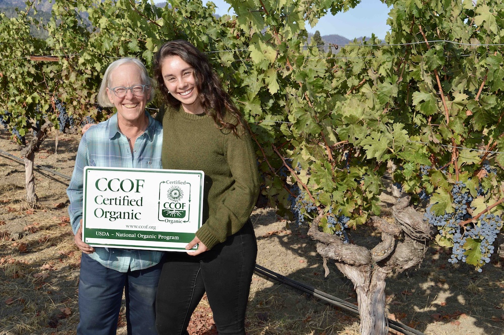 Cathy Corison with daughter Grace holding a sign saying "CCOF Certified Organic" in front of a Kronos grapevine.