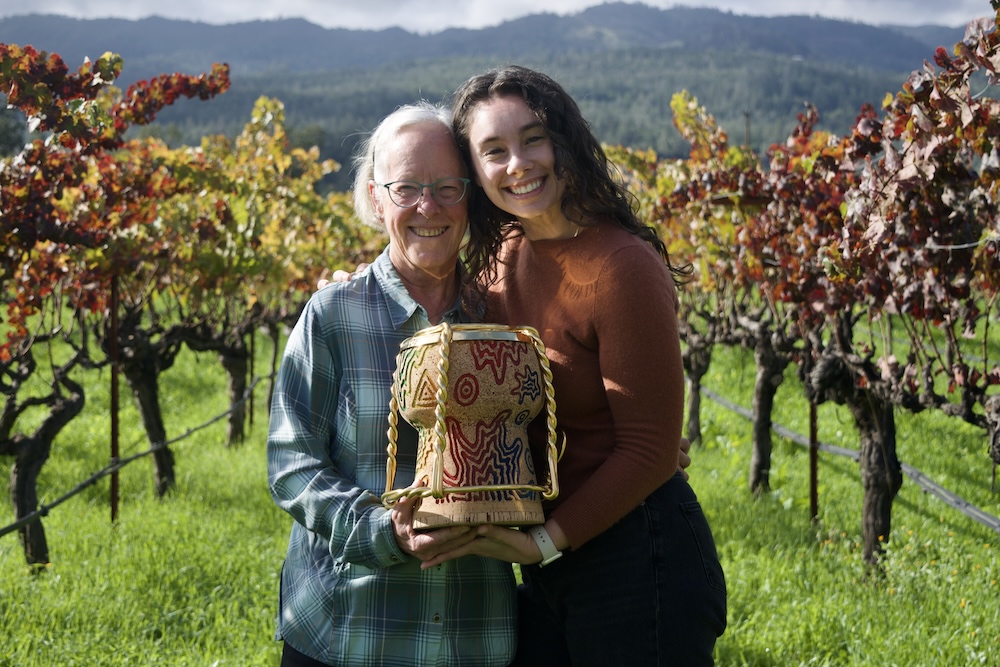 Cathy Corison and Grace Corison Martin holding the Golden Vines award, a giant decorated champagne cork, out in Kronos VIneyard.